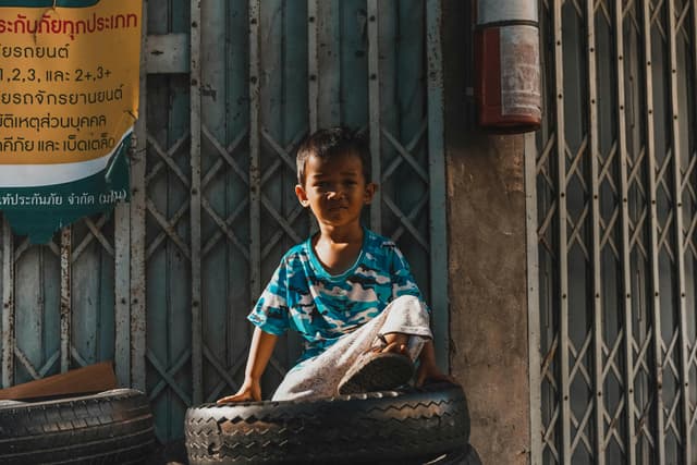 A young boy in a blue t-shirt sits on tires in a Bangkok street, Thailand.