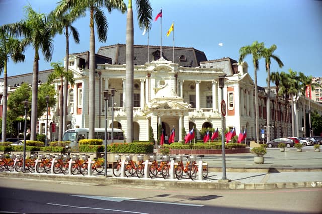 View of Taichung City Hall surrounded by palm trees and bicycles, under a clear blue sky.