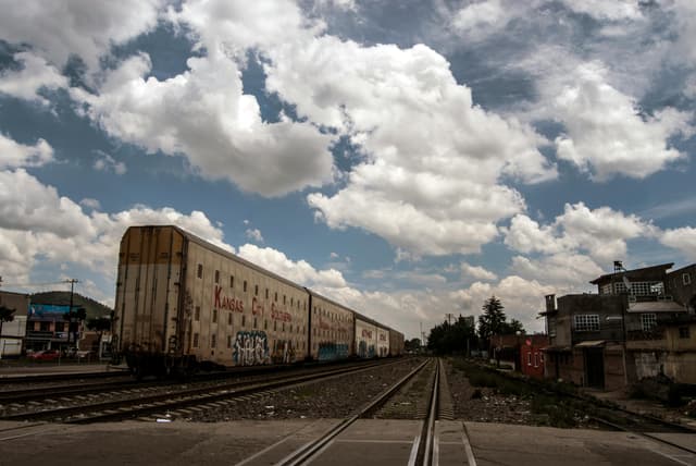 Freight train on tracks with cloudy sky and urban backdrop, captured during daytime.