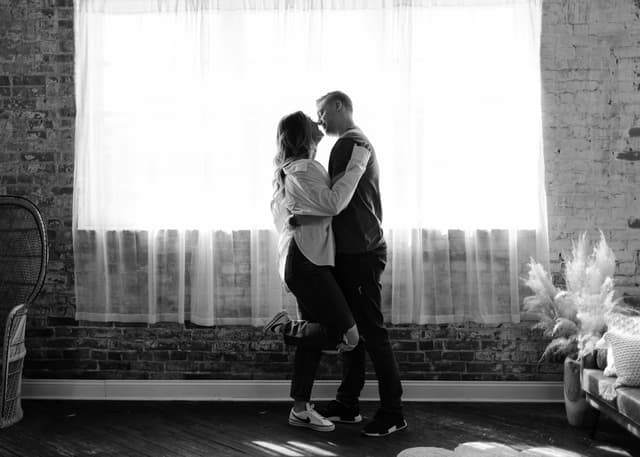A romantic moment of a couple embracing in a sunlit loft with brick walls and large windows.