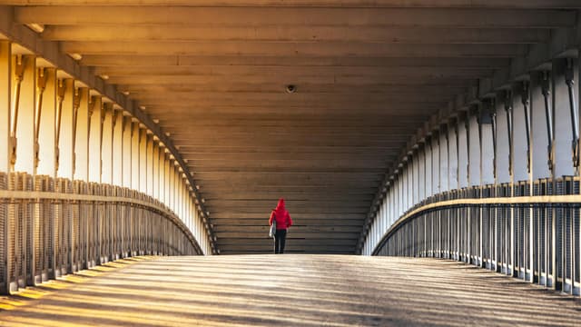 A lone figure in a red jacket walking under a bridge at sunset, casting long shadows.