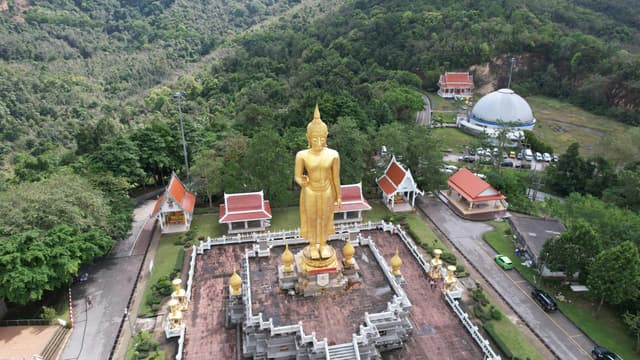 Aerial view of a golden Buddha statue in a temple complex amidst lush forest in Songkhla, Thailand.