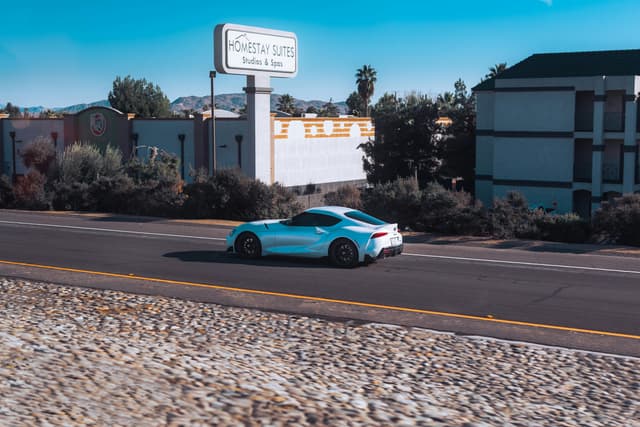 Blue Toyota GR Supra driving on road past Homestay Suites. Sunny day, clear sky.