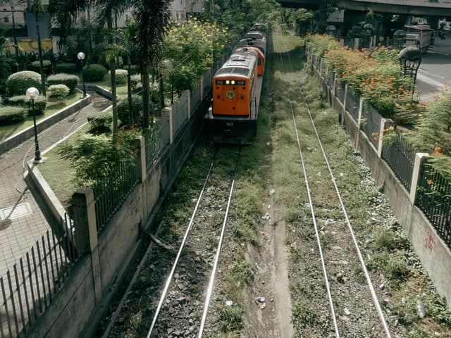 An orange train travels through lush greenery and urban structures in Makati, Philippines.