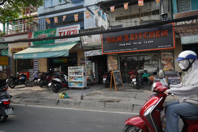 Bustling Vietnamese street scene featuring Dan's Kitchen cafe and passing motorbikes.