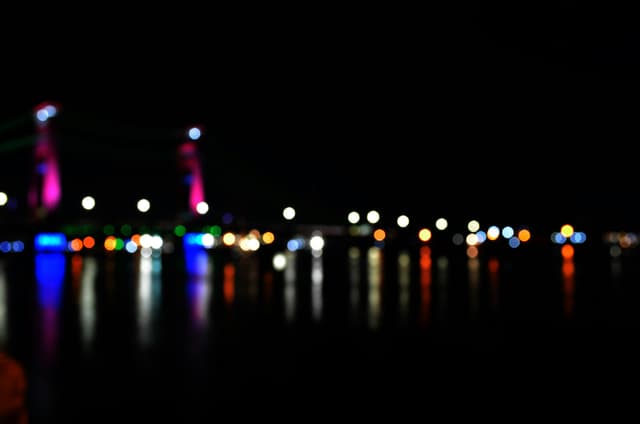 Blurred view of Ampera Bridge lights reflecting in water, creating bokeh effects.