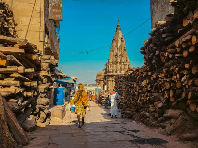 Street scene in Varanasi with wood piles and temple in view, showcasing daily life.