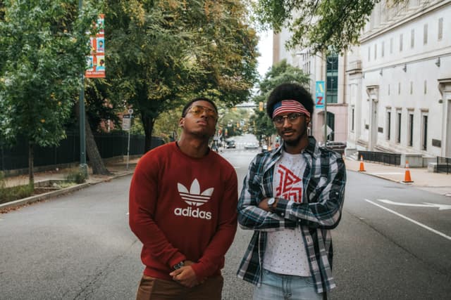 Two fashionable men posing outdoors on a city street with trees and buildings.