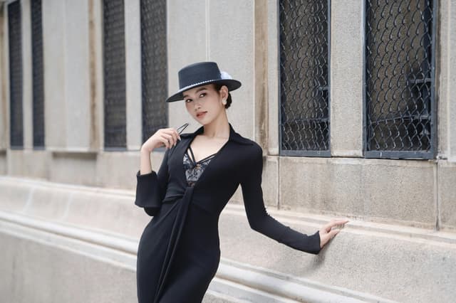 Stylish woman in black dress and hat poses against urban backdrop.