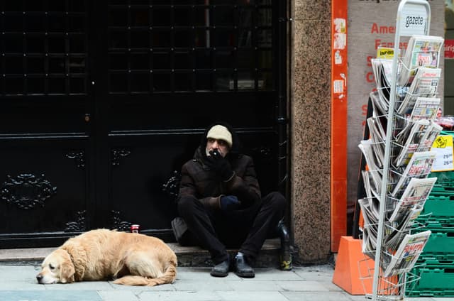 A man sits with his dog near a newsstand on a city street, capturing urban life.