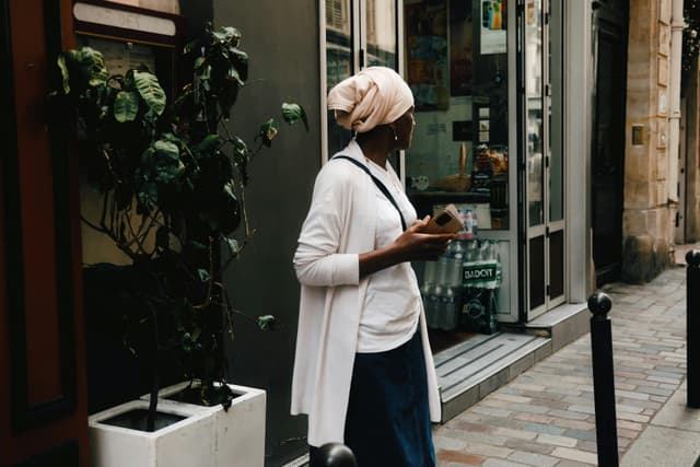 Stylish woman in a headscarf holding phone walking in a city street reflecting urban lifestyle.