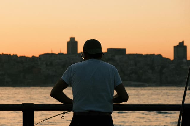 A serene silhouette of a man fishing by the water at sunset, with city skyline in the background.