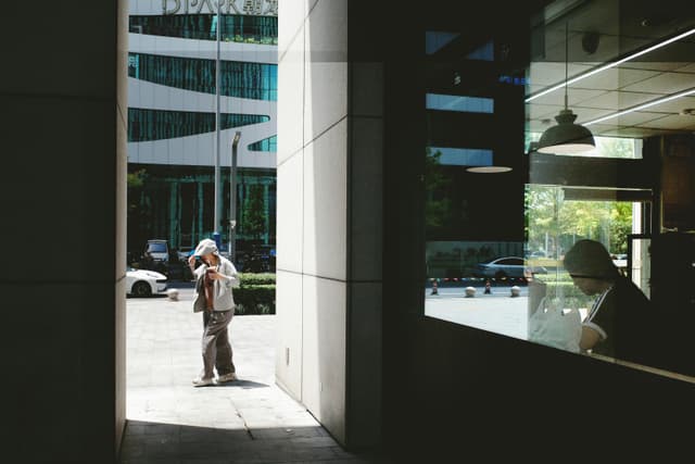 A person walking outside through a shadowed archway with reflections in a storefront window.
