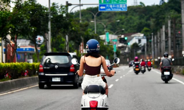 A woman rides a scooter through a busy city street, surrounded by traffic.