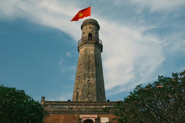 A scenic view of a historic lighthouse in Nam Định, Vietnam, crowned with the Vietnamese flag.