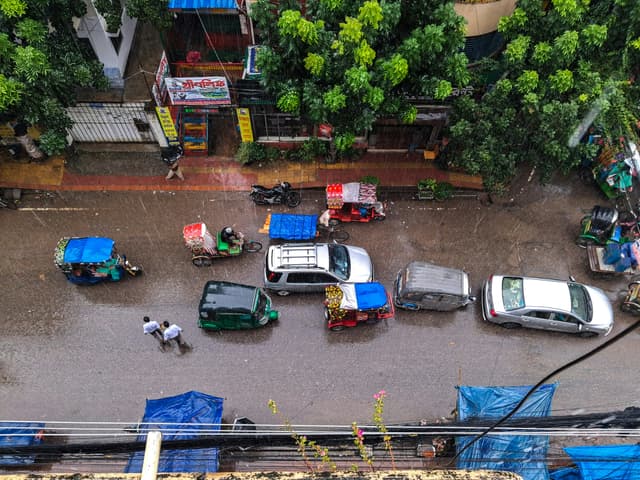 Aerial view of a Dhaka street with vehicles and pedestrians in the rain.