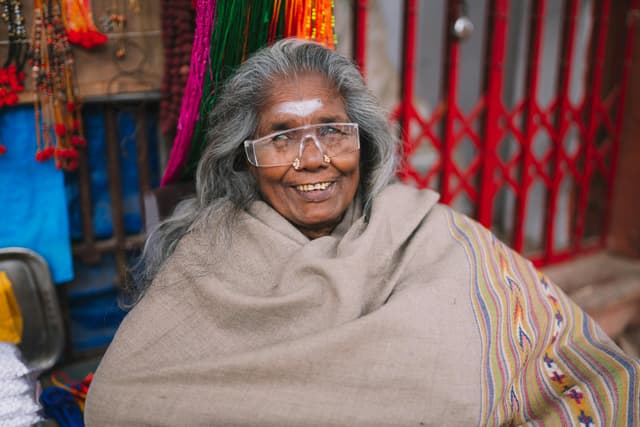 Elderly woman with glasses and traditional shawl smiling at a Varanasi market stall.
