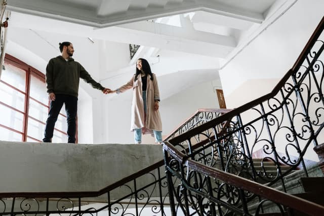 A couple romantically holds hands while standing on an ornate staircase indoors, exuding a warm and classic vibe.