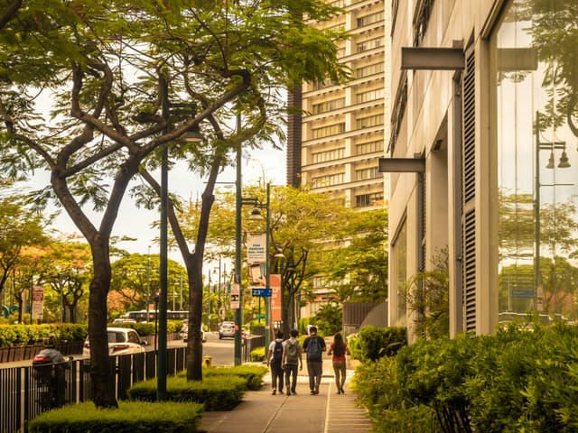 People walking on a tree-lined sidewalk in a bustling Taguig street, surrounded by modern architecture.