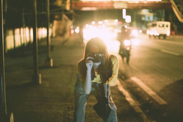 A woman bends forward on a street at night with an analog camera, capturing the city's ambiance.