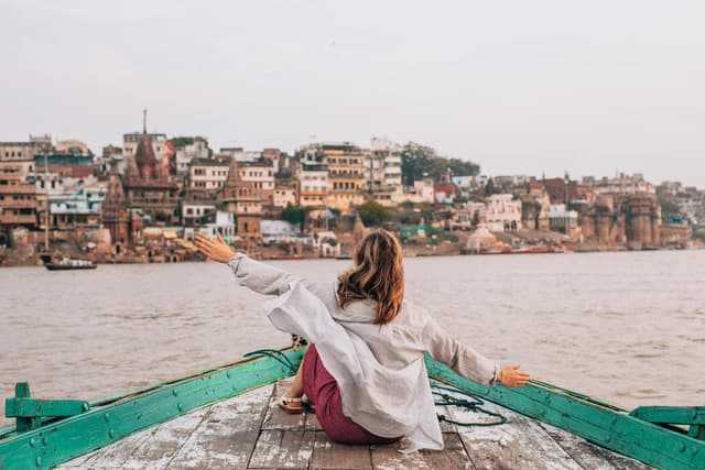 Woman sitting on a boat with arms raised, overlooking the ghats of Varanasi, India.
