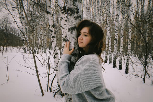 A woman embraces a birch tree in a snowy forest, capturing a serene winter moment.