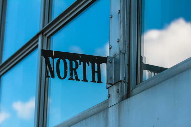 Reflective glass building corner with metal sign reading 'North' against blue sky and clouds.